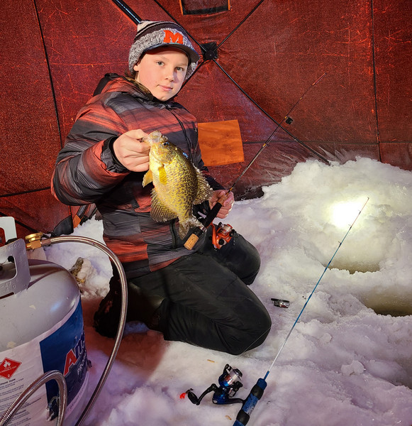 A boy joyfully holds a fish in the snow, showcasing his triumph in catching it amidst the wintry landscape.