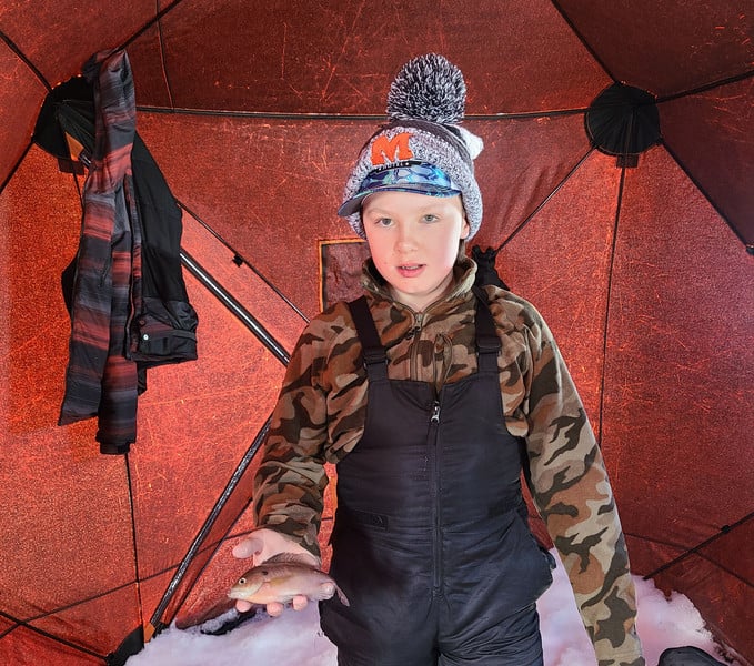 A young boy in a hat and overalls standing inside a tent.