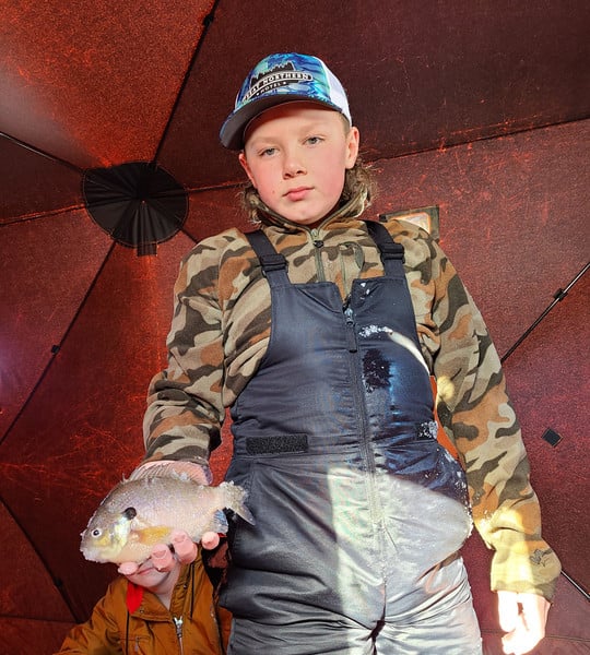 A young boy wearing a hat and overalls joyfully holds a fish in his hands.