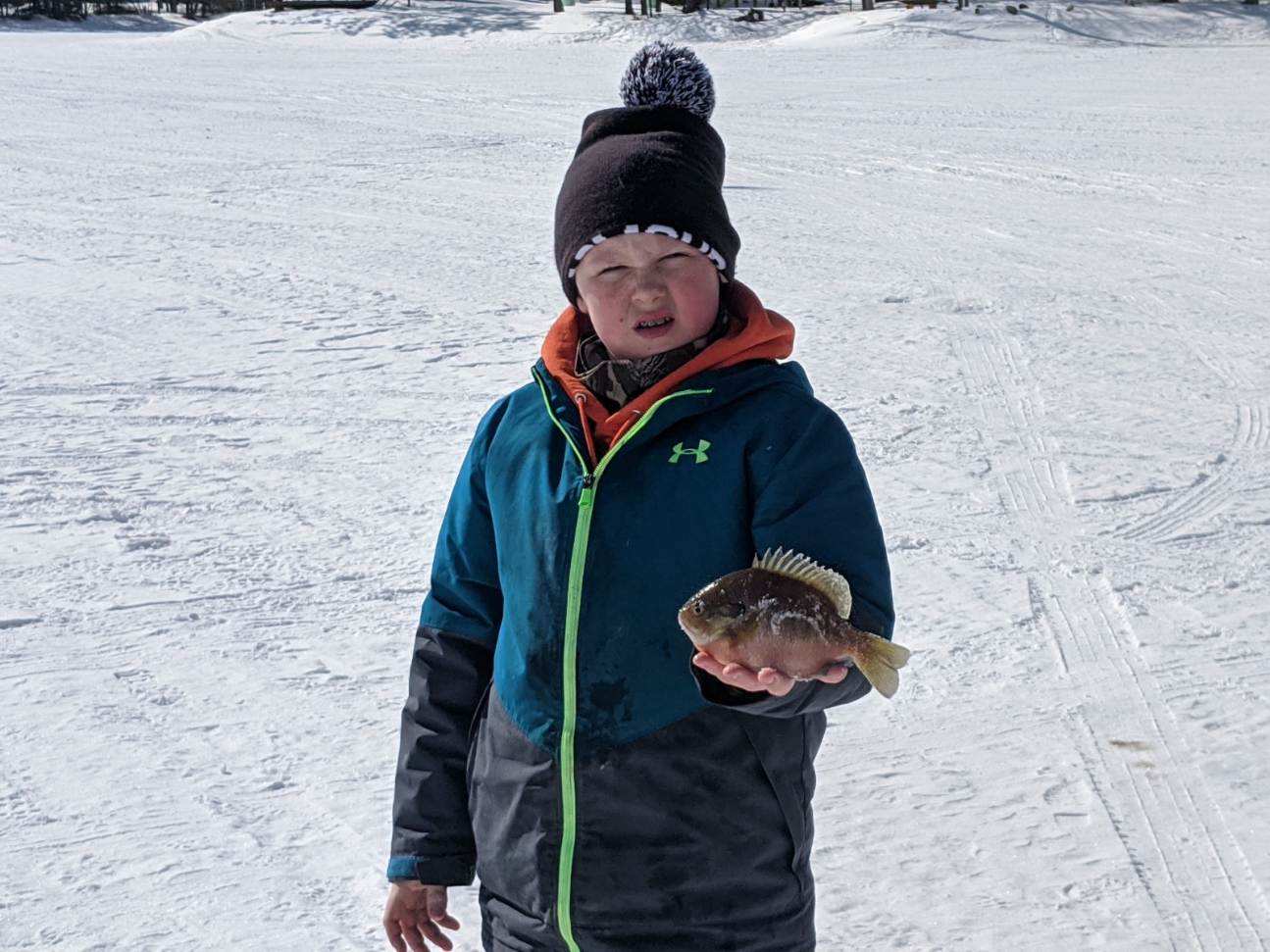 A young boy holding a fish in the snow, smiling brightly.