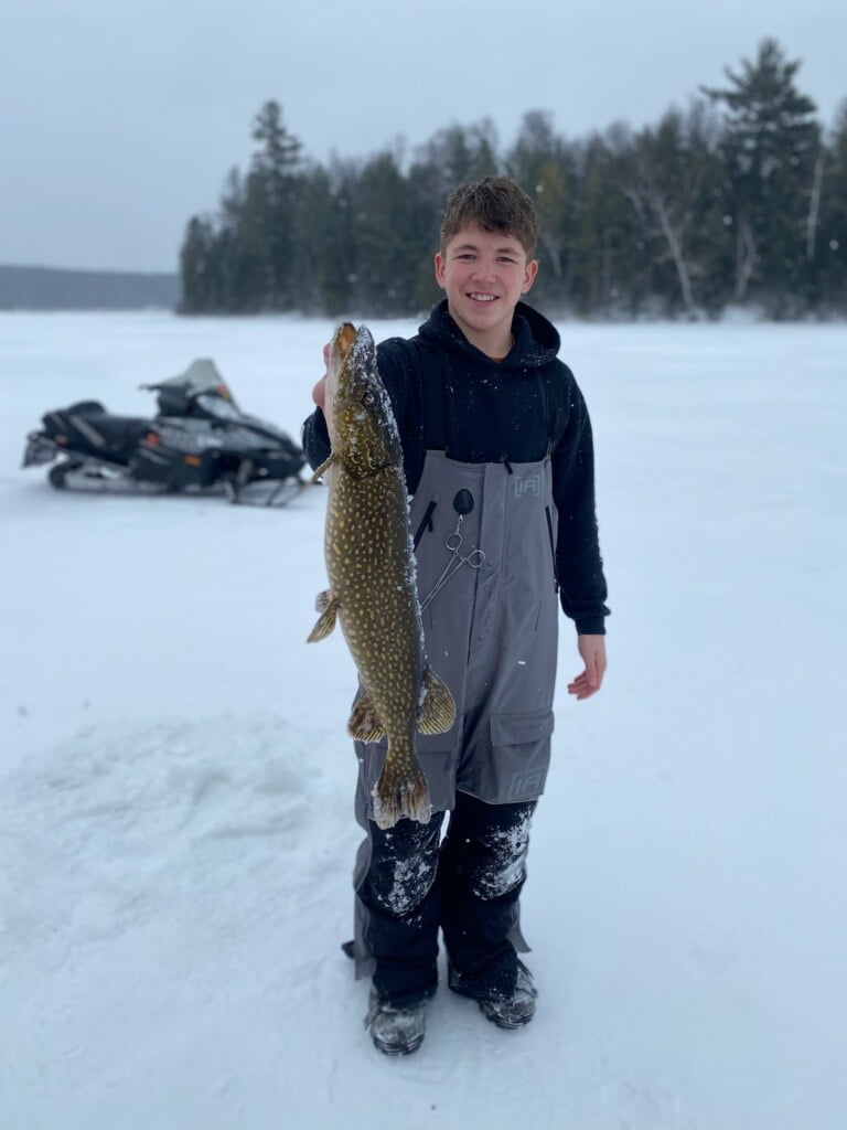 A boy proudly holds a large pike fish he caught on a snowy lake, showcasing his fishing skills.