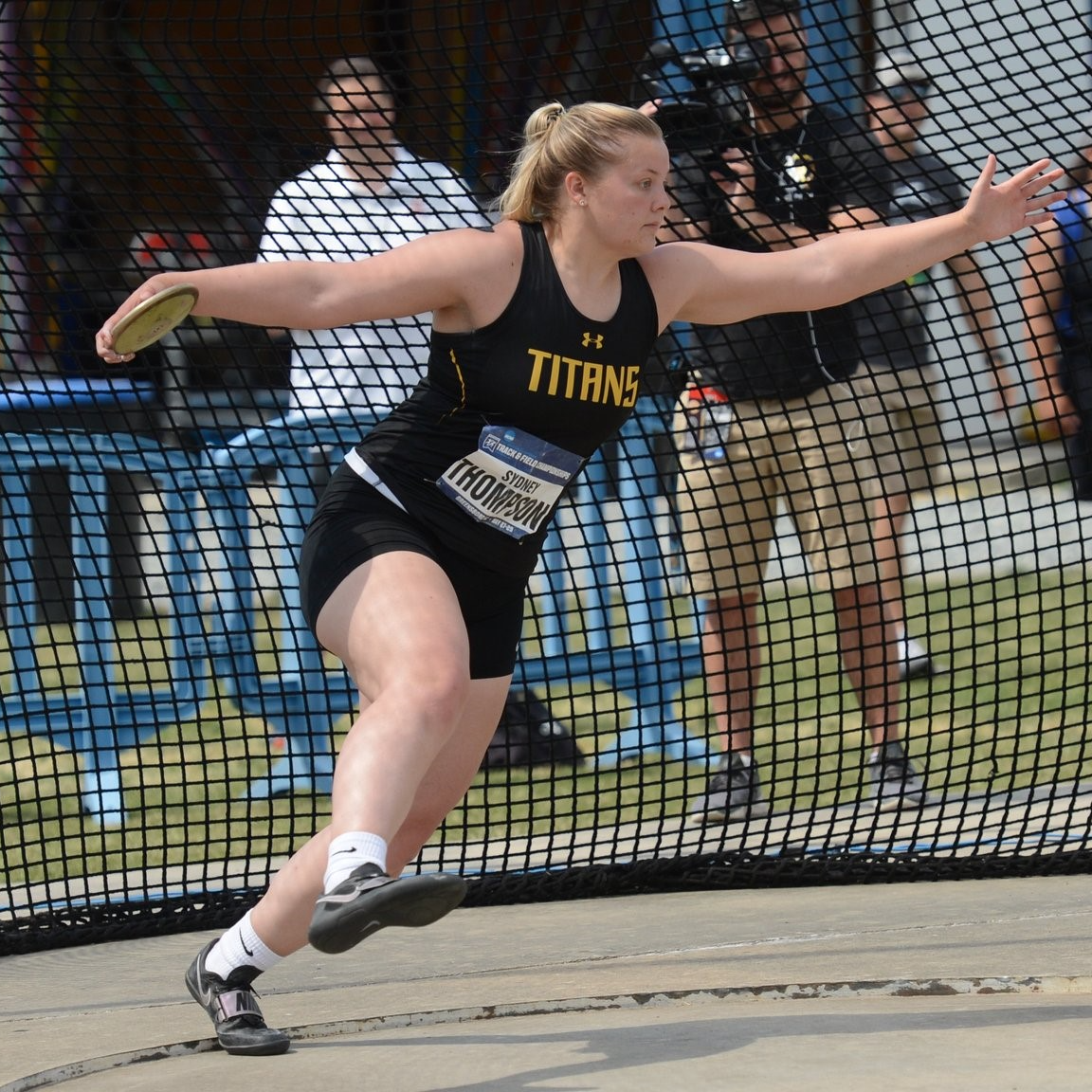 Woman, with name Sydney Thompson on front, in Titans track jersey throwing discus . In background is a cameraman an spectators