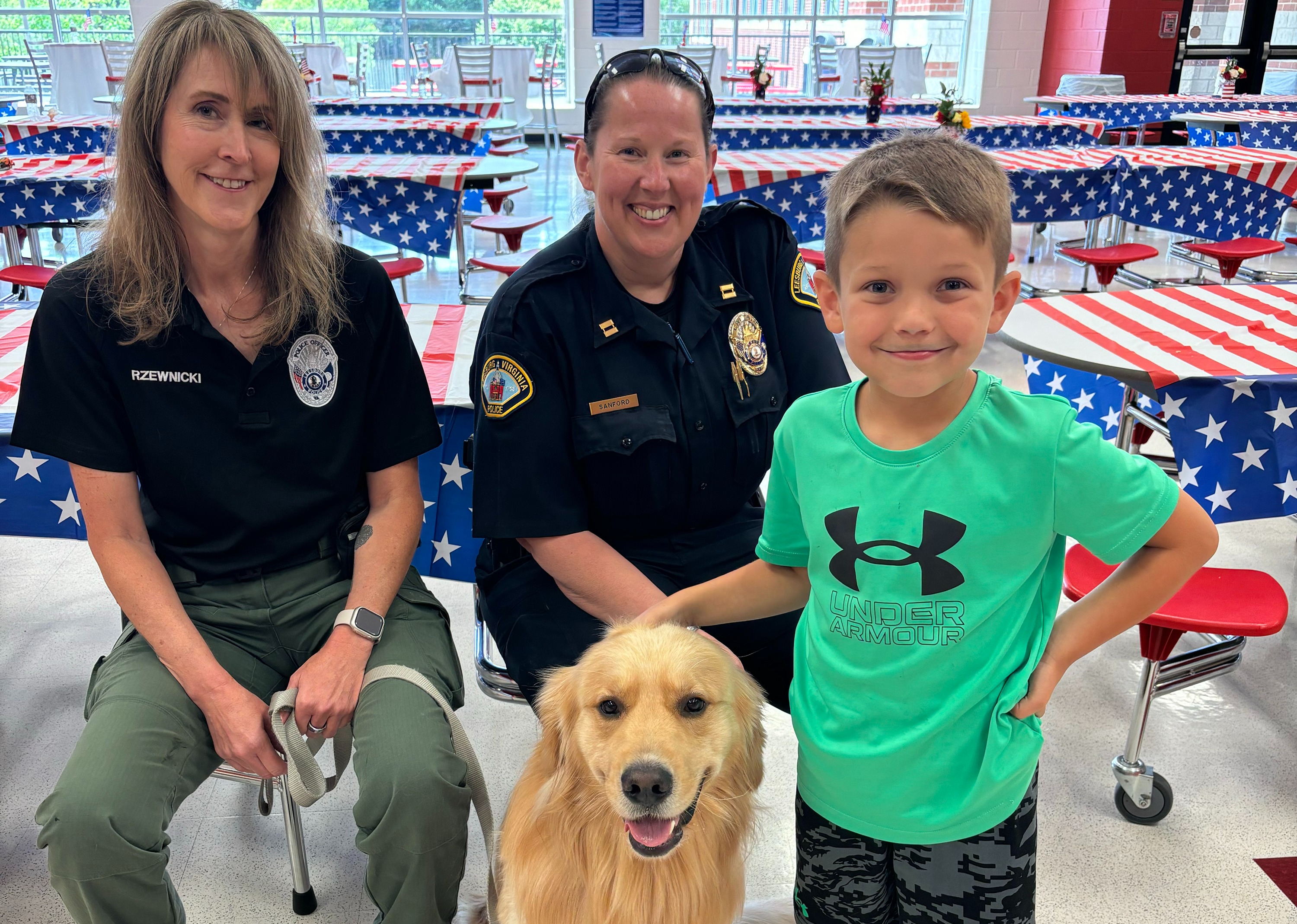 Two officers and a young boy smile for a photo with a golden retriever sitting between them. The background shows tables covered in American flag-themed tablecloths in a bright cafeteria setting.