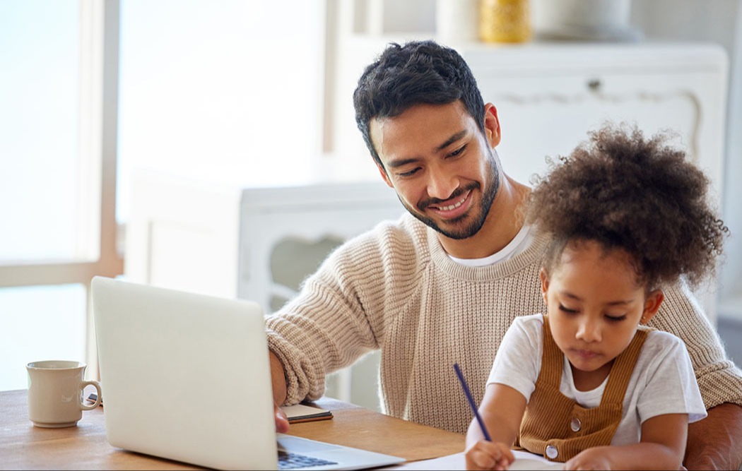 Parent assisting young child with homework at a table, using a laptop.