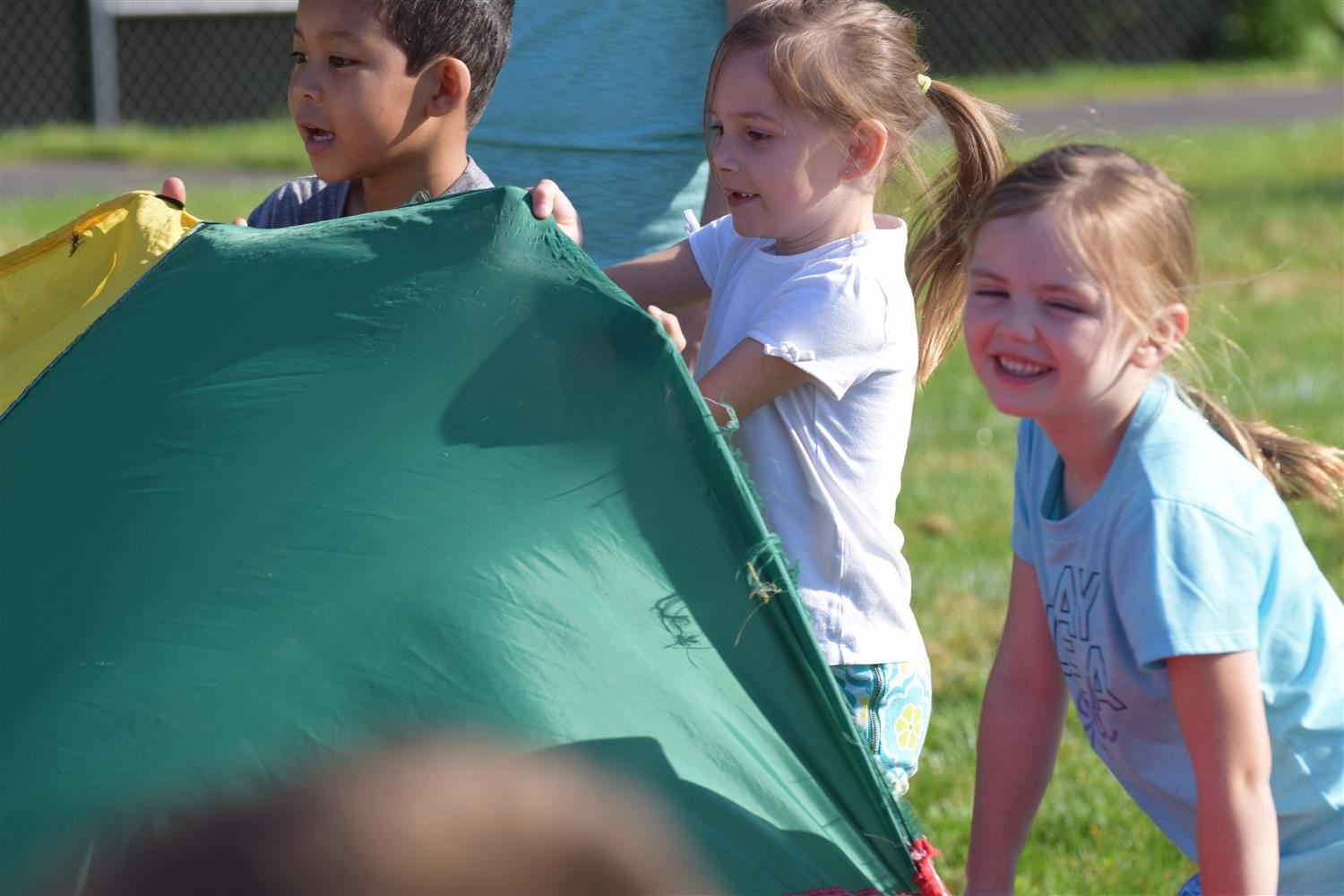 Three young children playing with a large, colorful parachute outdoors on a sunny day, smiling and engaged in the activity.