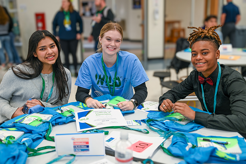 Three students sitting at a table with t-shirts and lanyards on it.