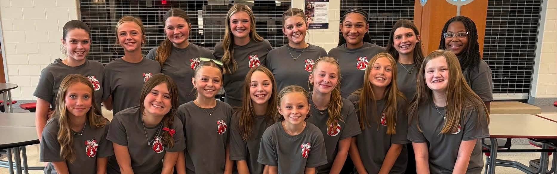 A group of young girls in matching gray shirts pose for a picture in a cafeteria.