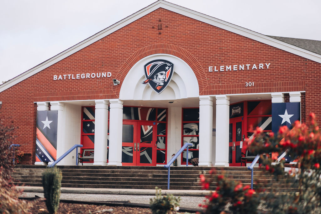 A red brick school building with white trim features the words "Battleground Elementary" above the entrance.