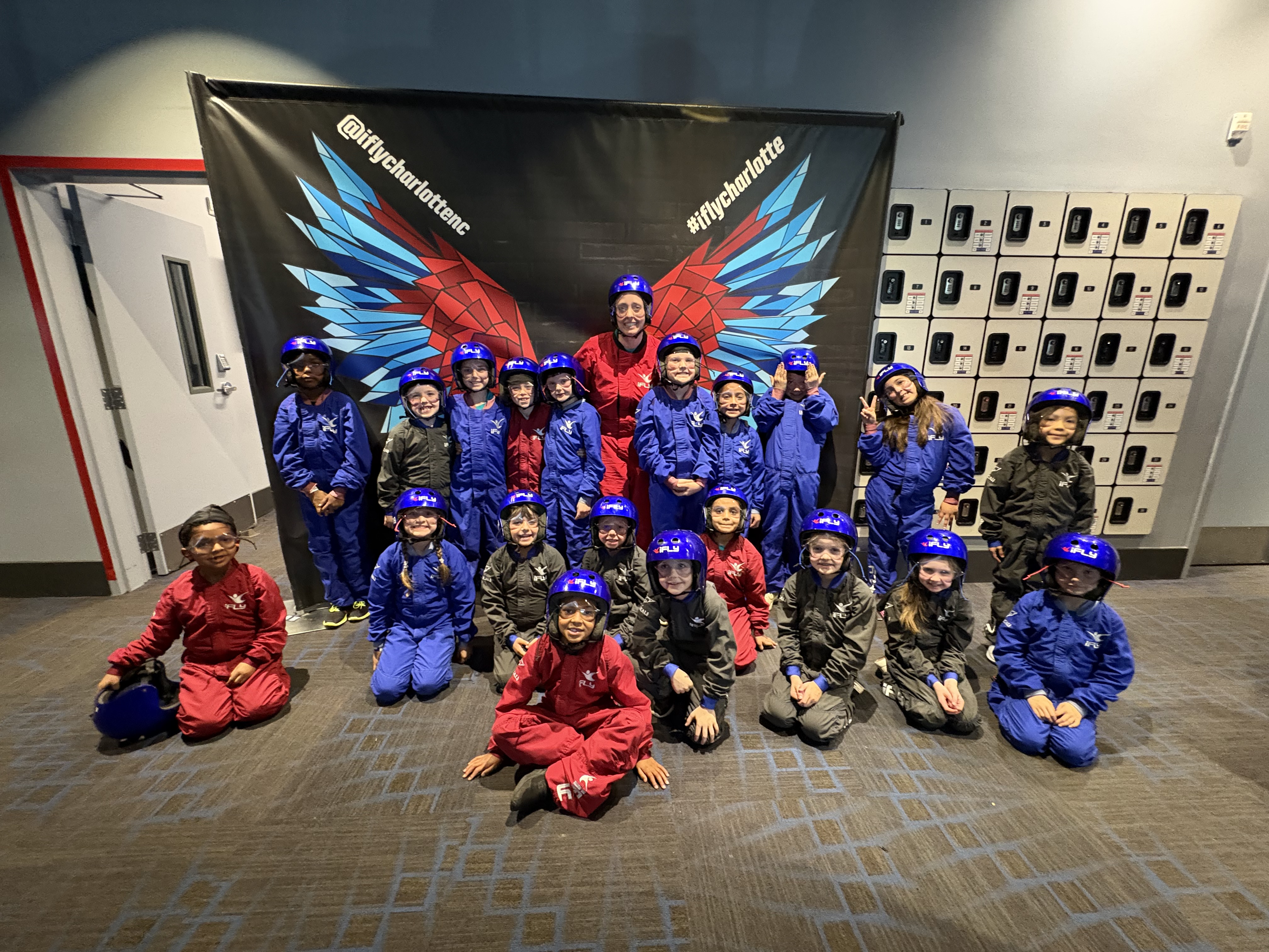 A large group of elementary school students and one adult instructor pose for a group photo at iFLY Charlotte. The students are wearing indoor skydiving flight suits in shades of blue, red, and charcoal gray, along with blue safety helmets and goggles. They are positioned in front of a large black backdrop featuring a set of stylized red and blue wings, flanked by the social media handles @iflycharlottenc and #iflycharlotte. Some students are standing in the back row while others are kneeling or sitting in the front on a patterned gray carpet. To the right, a wall of white lockers is visible.
