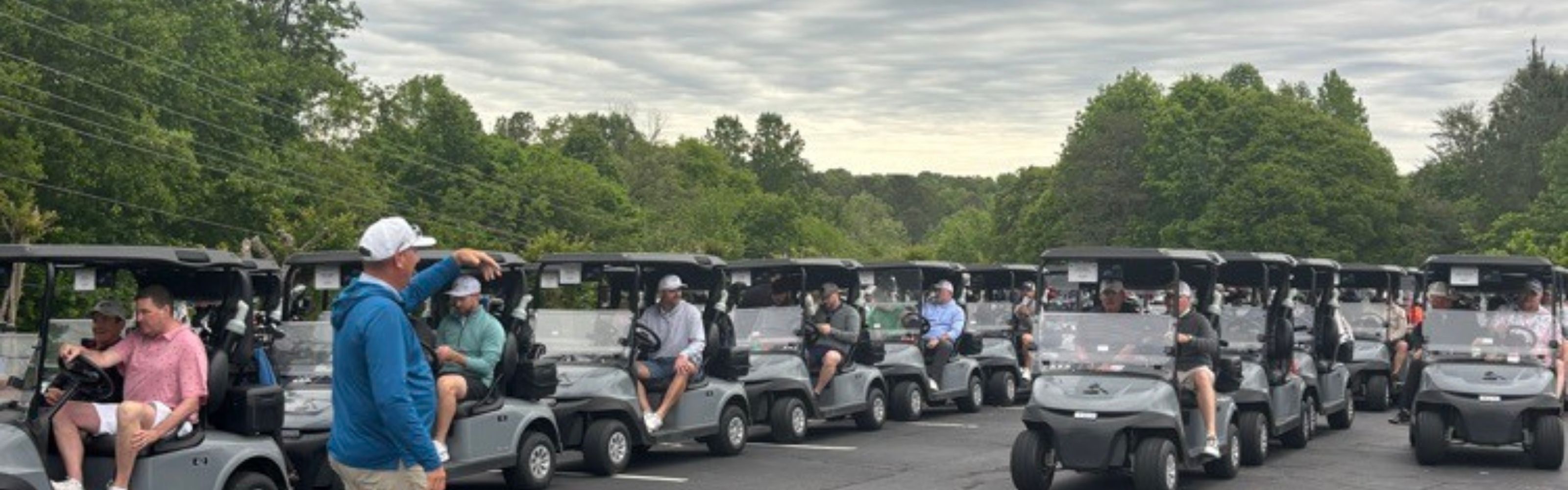 People in golf carts in a parking lot with a row of trees in the background.