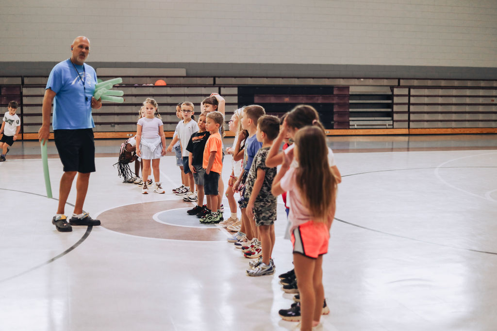 A man leads a group of children on an indoor basketball court. They stand in a line, listening.