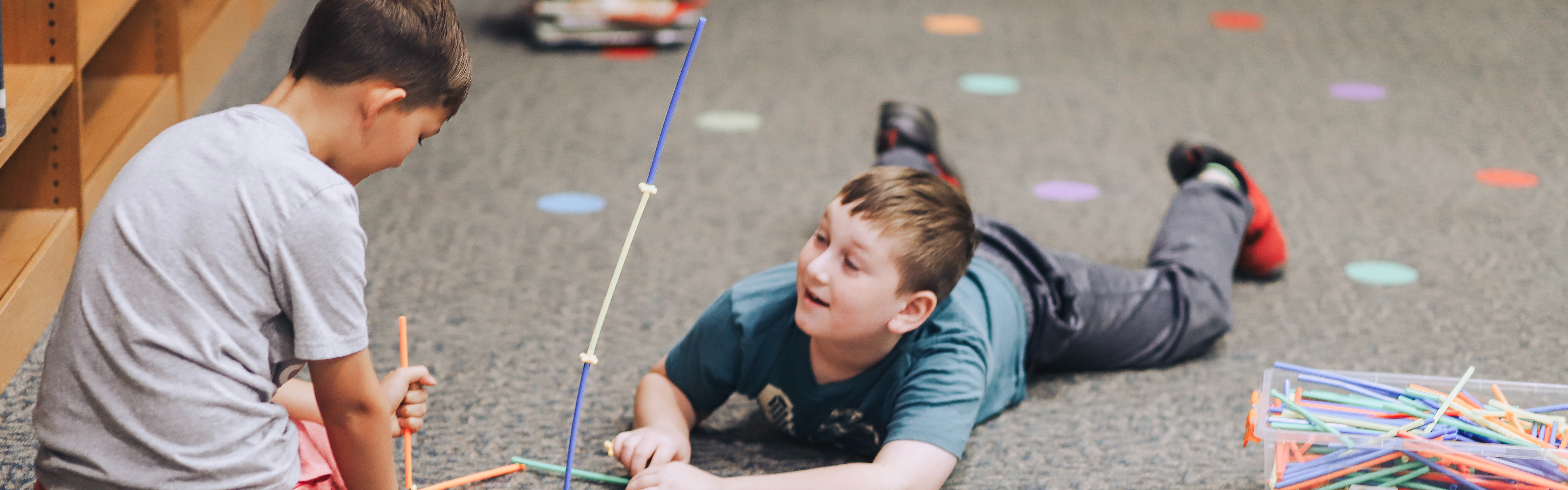 Two young boys play with a colorful straw and connector building set on a gray carpeted floor inside a classroom or library setting. One boy, wearing a grey t-shirt and pink shorts, is kneeling on the left and holding up a structure made of orange, green, and blue straws. The second boy, wearing a teal t-shirt and grey pants, is lying on his stomach on the right, smiling and looking up at the first boy as they work together. A clear plastic bin filled with building straws sits on the carpet next to a clear plastic lid, and bookshelves are visible in the blurred background.
