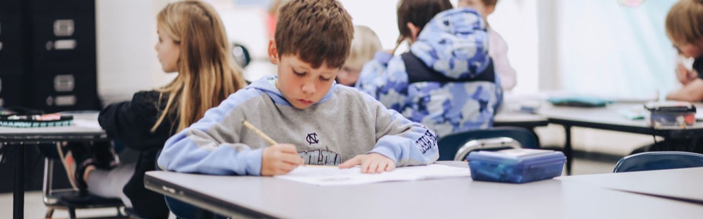 Kids sitting at desks in a classroom. One boy in front writing on paper with a pencil.