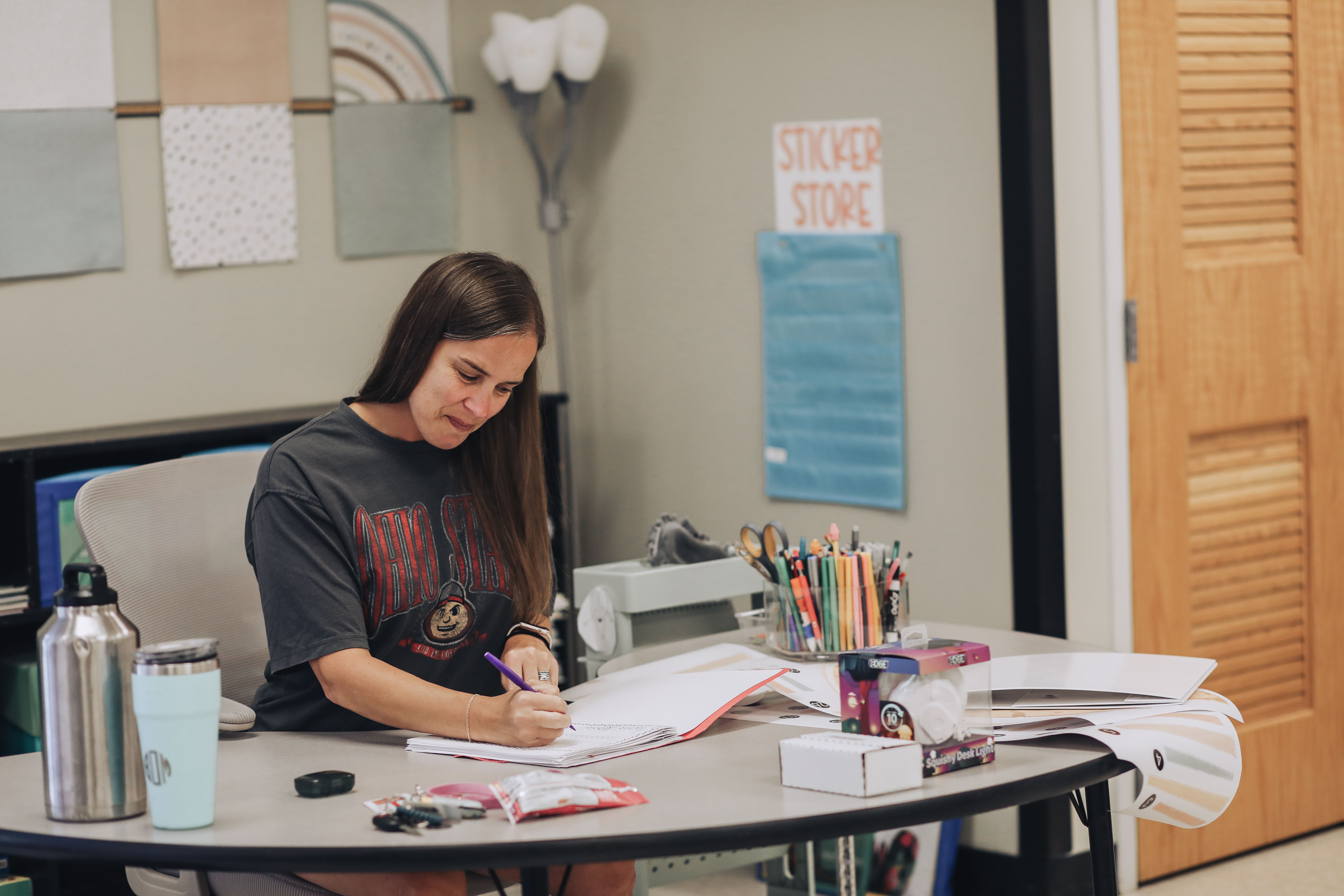 A teacher wearing a dark gray Ohio State shirt sits at a rounded desk in a classroom, writing in a open binder or notebook with a purple pen. On the desk are water bottles, school supplies, classroom materials, and packaging. In the background, a sign on a wall pocket chart reads STICKER STORE.