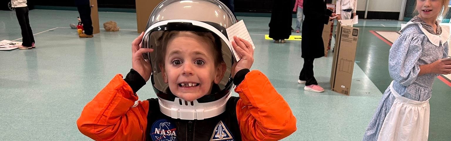 A child in a space suit stands in front of a sign for John Glenn.