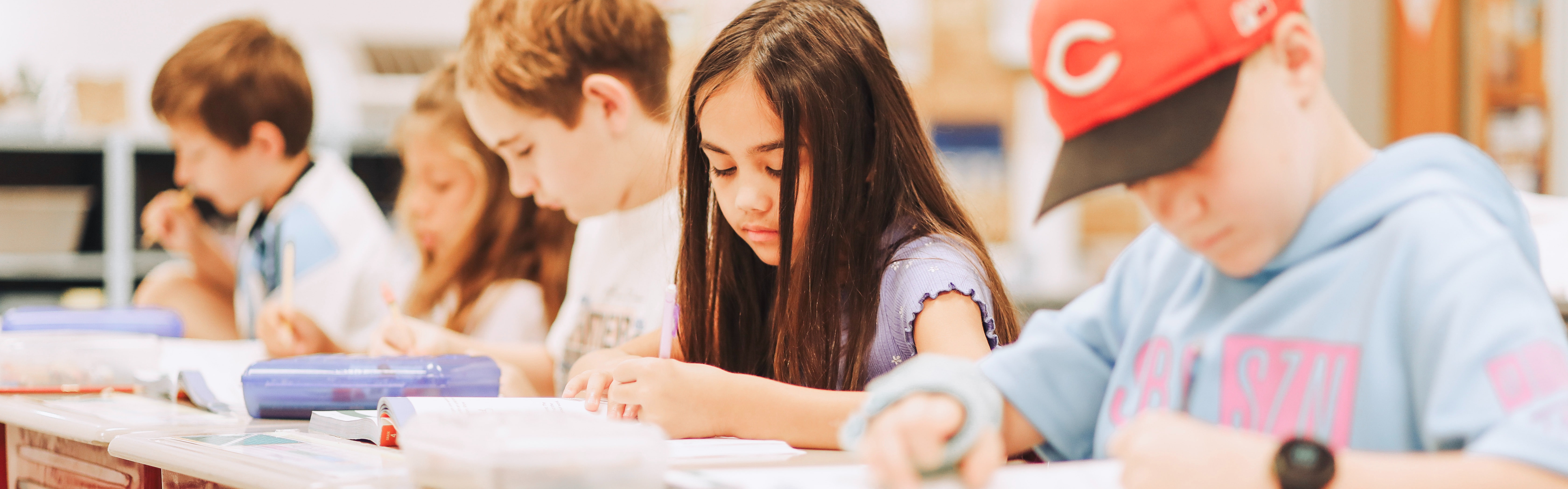 Kids seated in a classroom, writing on papers, with one wearing a red baseball cap.