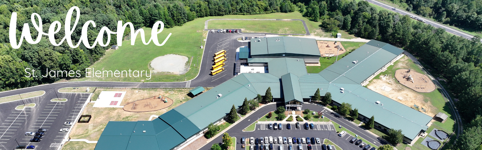 Aerial view of a school with green roofs, a parking lot, playground, and trees. The word "Welcome" is displayed.