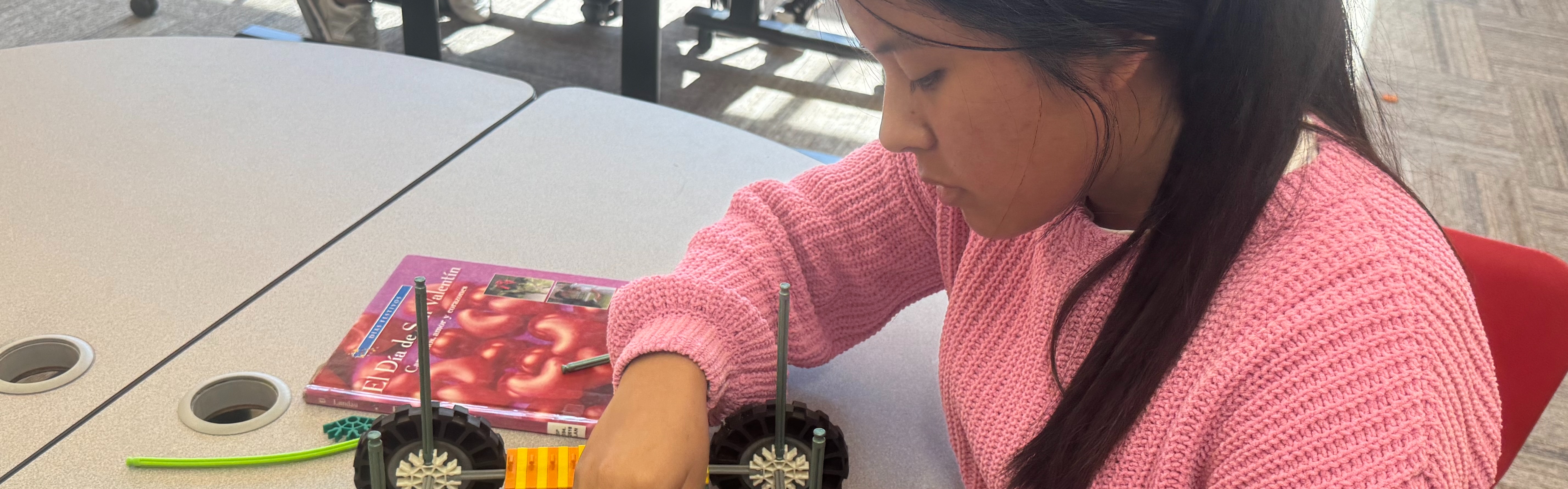 A close-up shot of a student wearing a pink sweater sitting at a light gray table. She is using both hands to assemble a complex wheeled vehicle made of K'Nex building pieces. Various green, orange, and red connectors are scattered on the table next to a red book.
