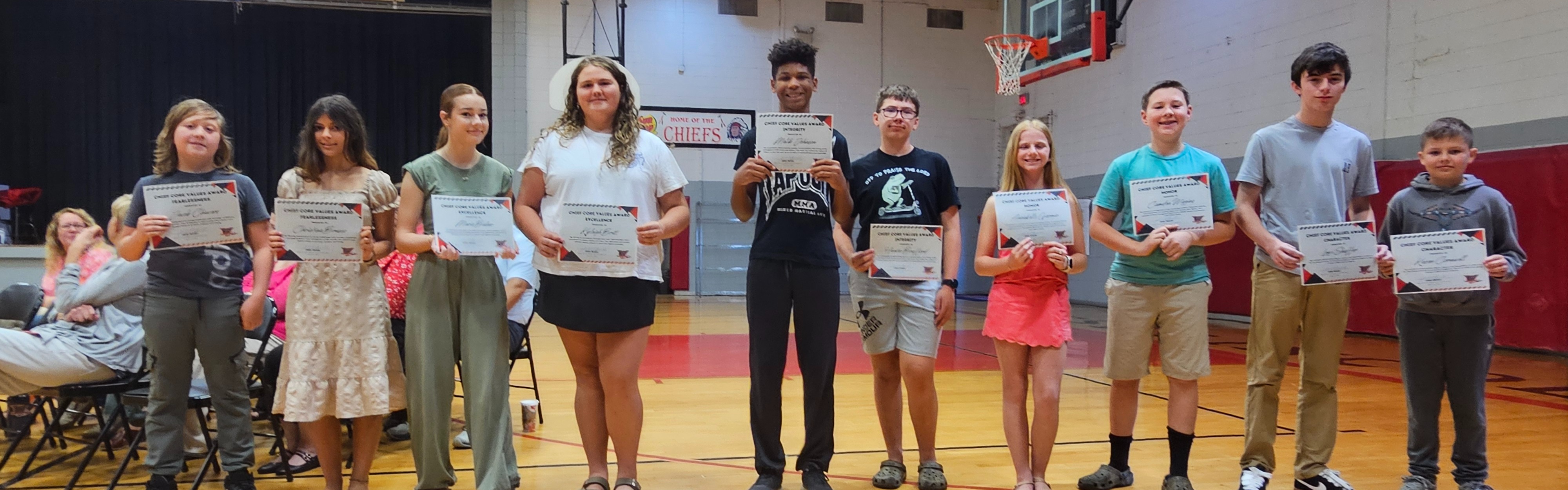 A group of people stands in a gymnasium holding certificates. Wooden flooring and basketball hoops are visible in the background.