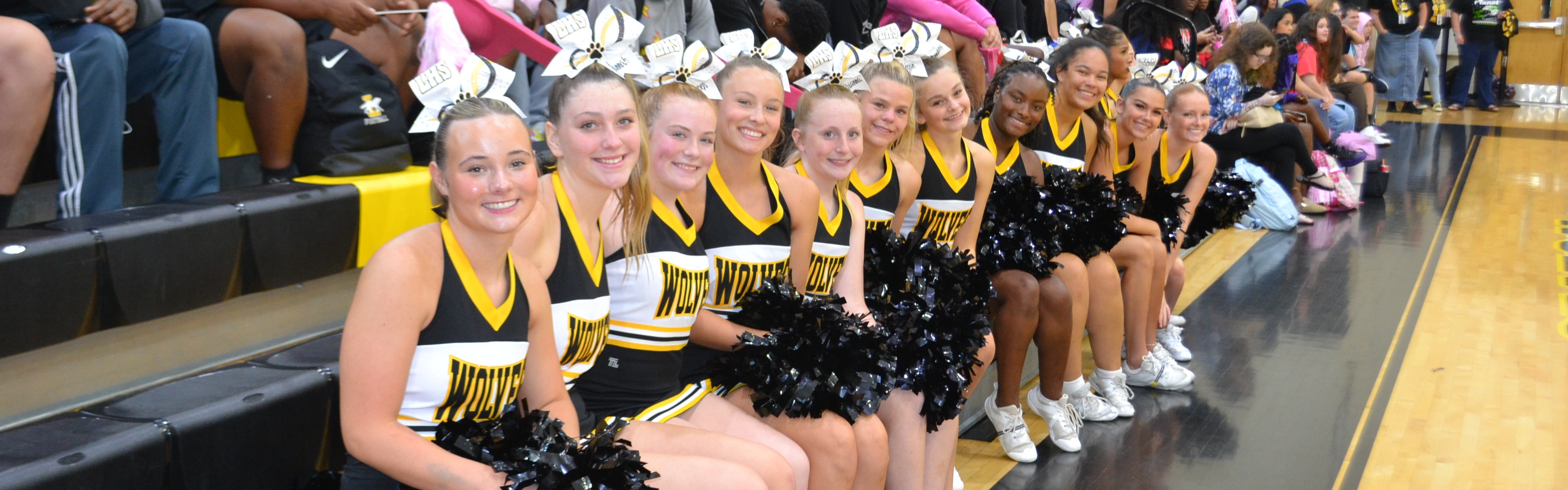 Cheerleaders sit on bleachers in a gym at a pep rally. A crowd of people sits behind them. Floor has black and brown wood.