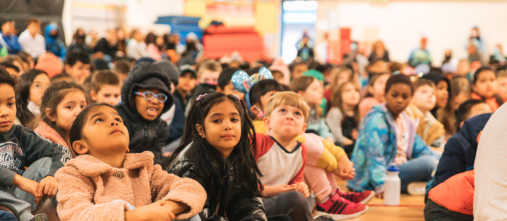 A group of children sit on a gym floor attentively watching something in front of them