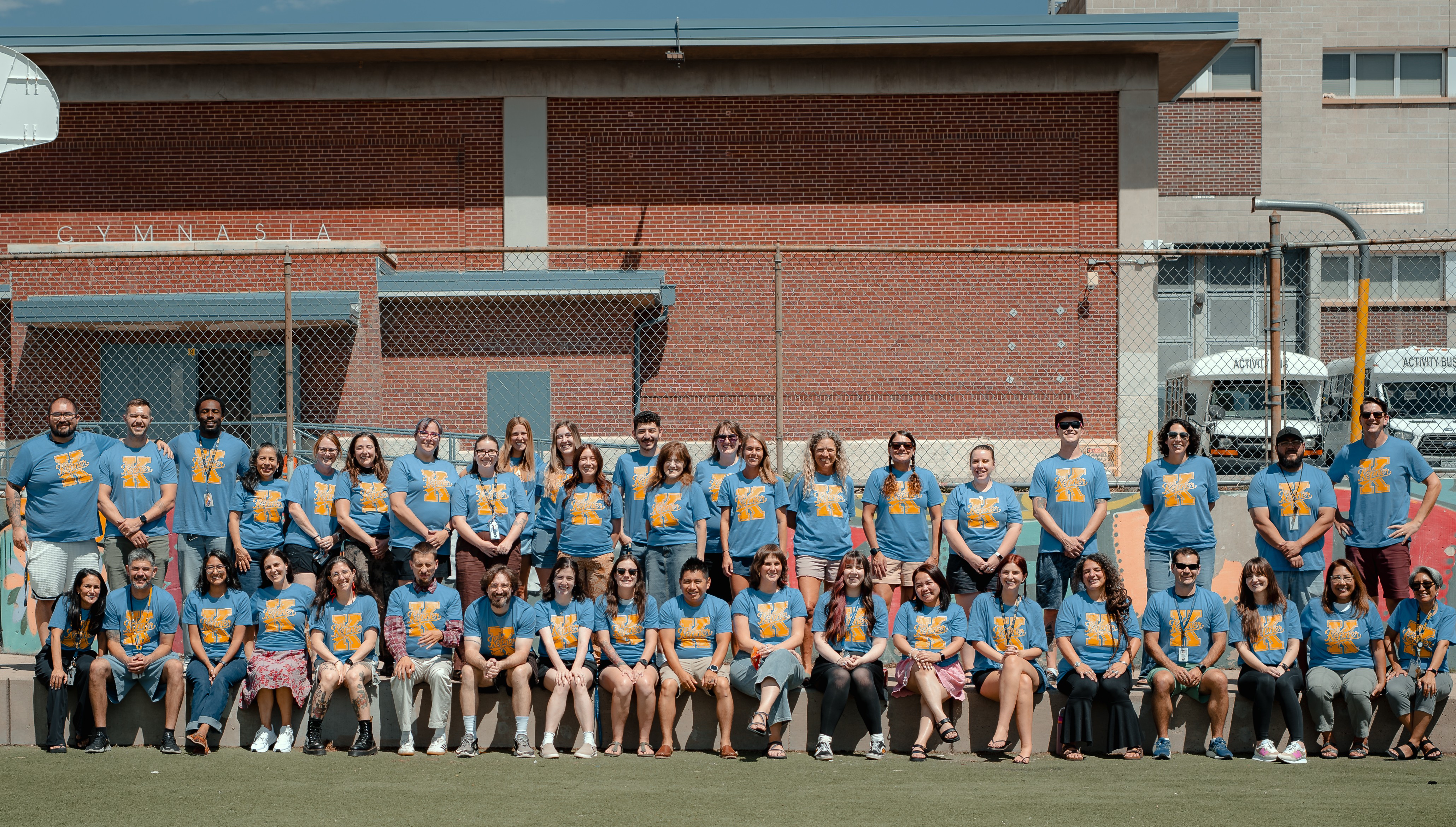 A group of people in blue shirts stands in a line in front of a brick building.