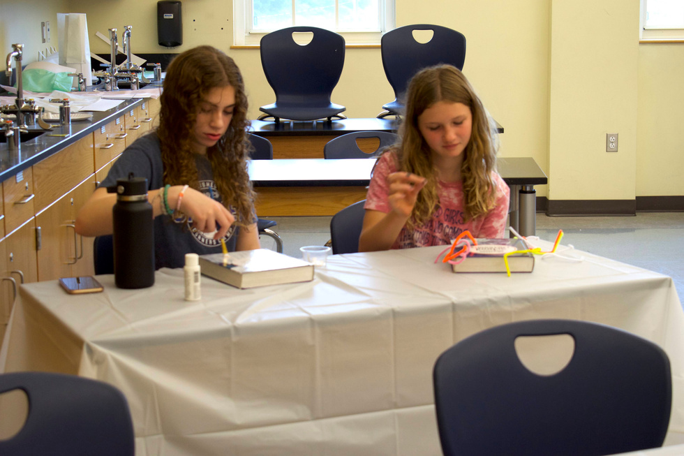 Two young girls sitting at desks in a classroom, with one of them working on a craft project.