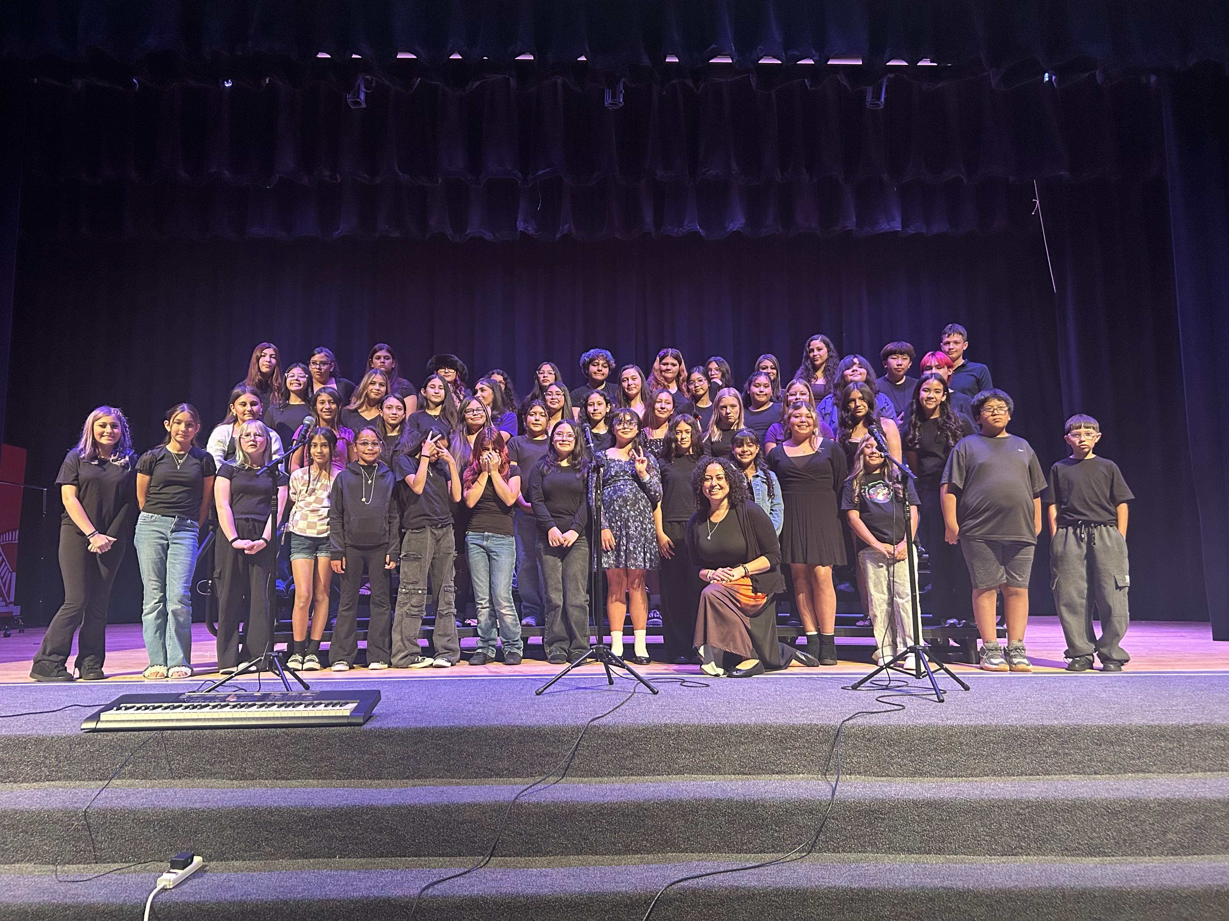 Many students and adults stand on stage in front of a dark backdrop with musical instruments on stands.