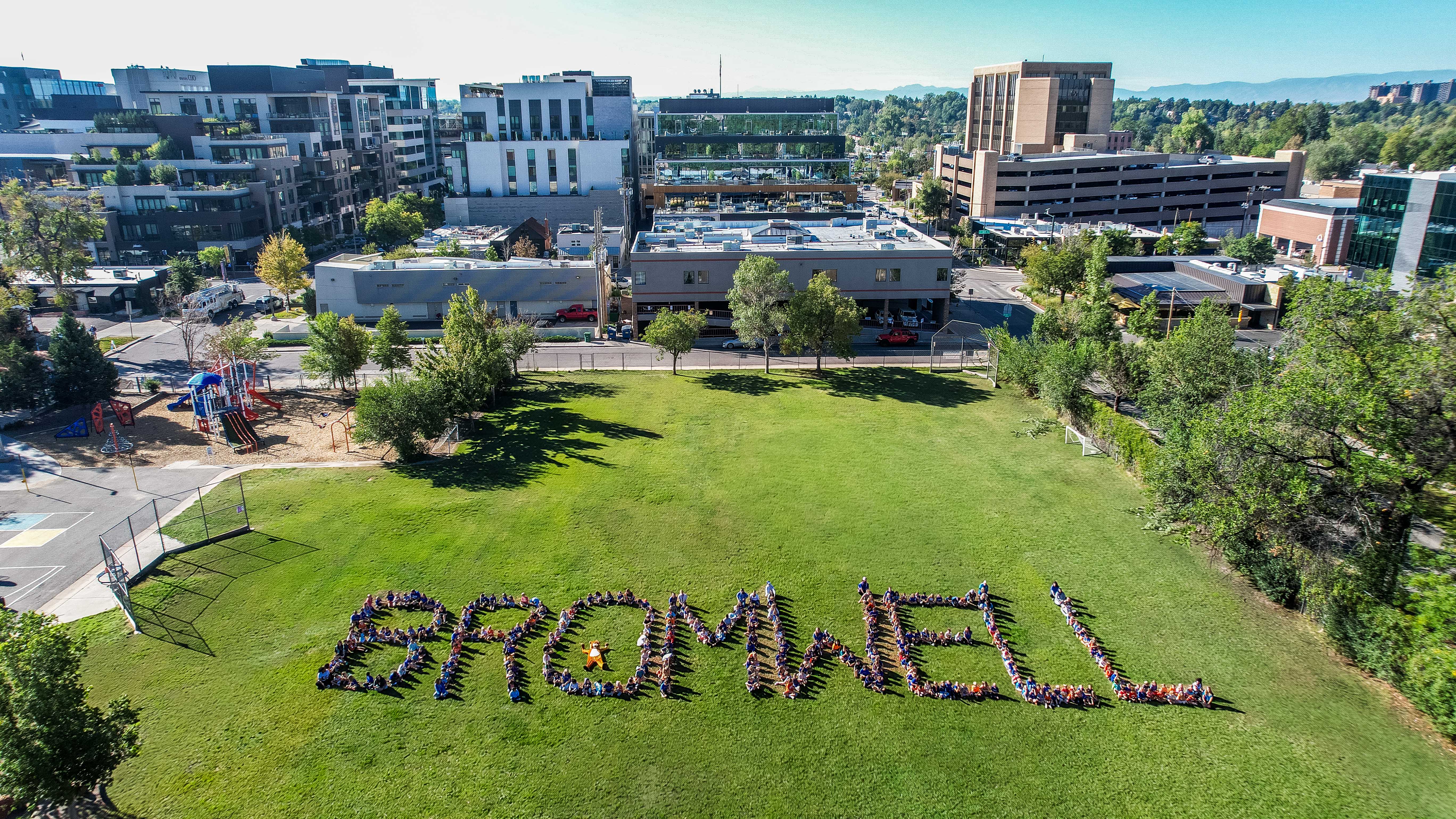 Aerial view of a large grassy field with the word "BROMWELL" spelled out in people. Cityscape with buildings.