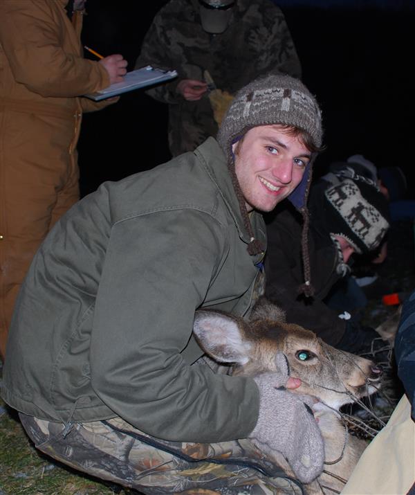 A guy wearing a green jacket and a hat while holding a deer with guys writing in a paper at the back.