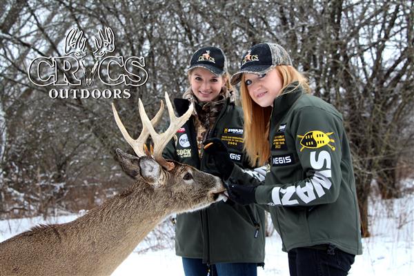 Two ladies both of them wearing green jacket and a cap, with reindeer in front of them.