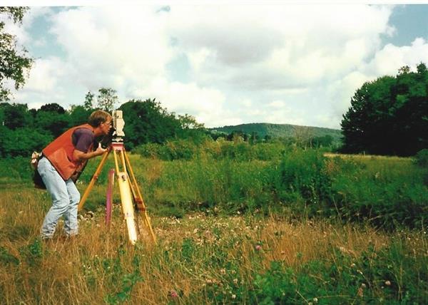 A guy wearing a red jacket trying to capture images with his camera in a greenfield.