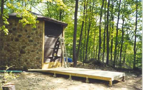 A wooden deck with a staircase leading up to a stone building with a gray slate roof. The building has small windows and a chimney. There are trees in the background.