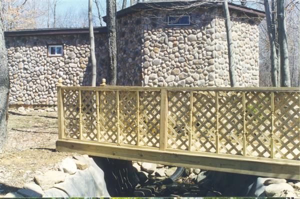 A wooden bridge with a railing leads across a stream to a stone house nestled amongst trees.