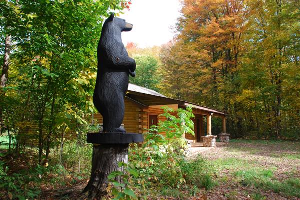 A statue of a black bear cub standing on its hind legs on a tree stump in front of a log cabin.