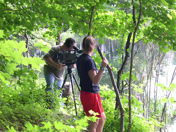 A lady and a guy taking a photo with his camera in the middle of a forest.