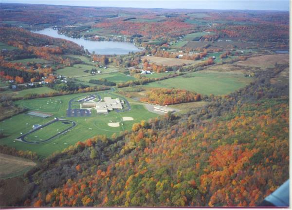 An aerial view of a red brick school building with a black roof surrounded by colorful trees in the fall.