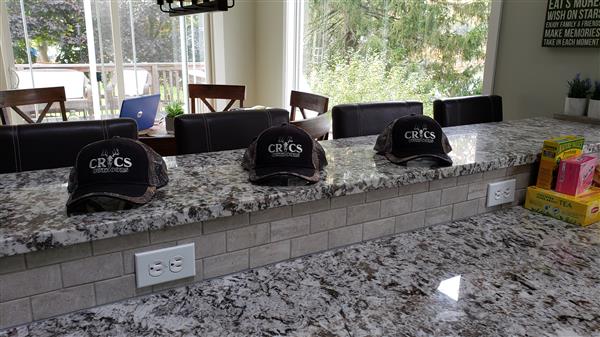 Three hats resting on a light colored kitchen counter. From left to right, a straw cowboy hat, a black baseball cap with white text, and a red knitted beanie with a white bobble on top.