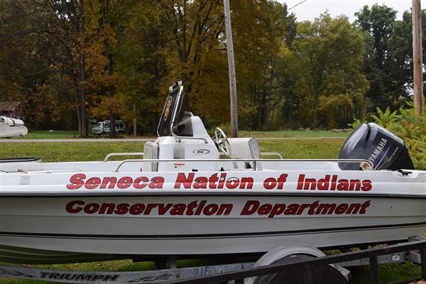 A white boat with the words "Seneca Nation of Indians Conservation Department" written on the side. The boat is parked on a trailer in a grassy field, with trees in the background.