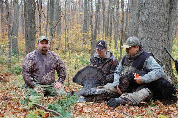 Three hunters wearing their full gear while holding a dog and a turkey in the middle of a forest.