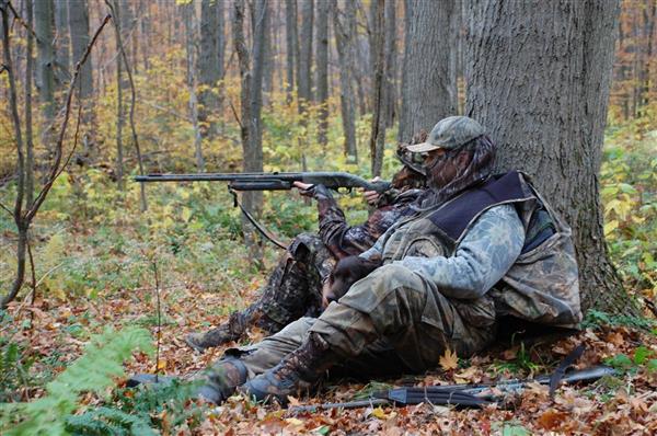 Two hunters wearing their full gear with their hunting guns and one of them is pointing the gun at something.