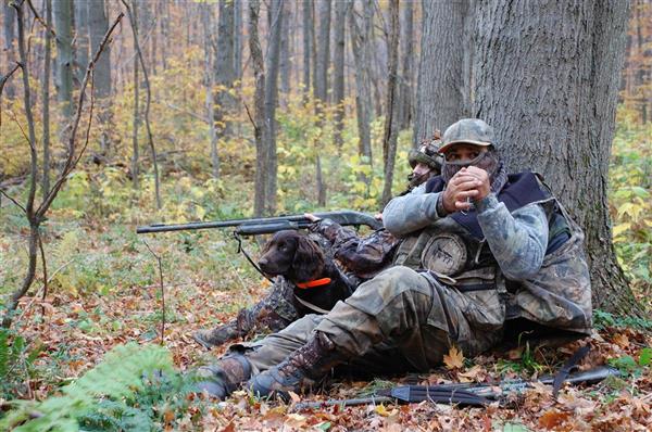 Two hunters wearing their full gear with their hunting gun posing for a picture while sitting under a tree.