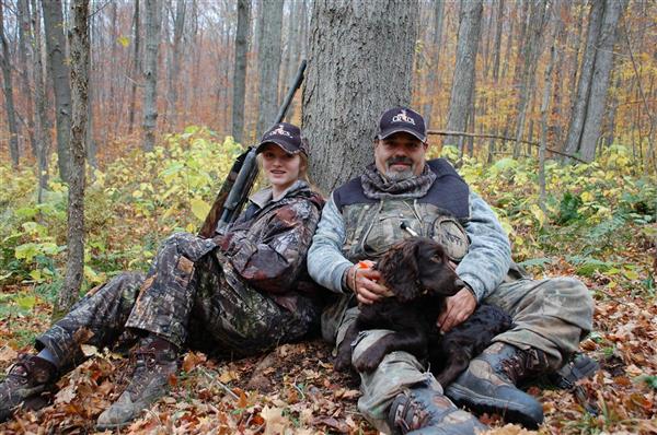 Two hunters wearing their full gear with their hunting guns sitting under a tree with their dog.