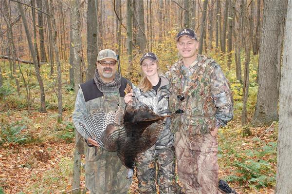 Three hunters wearing their full gear while holding a turkey.