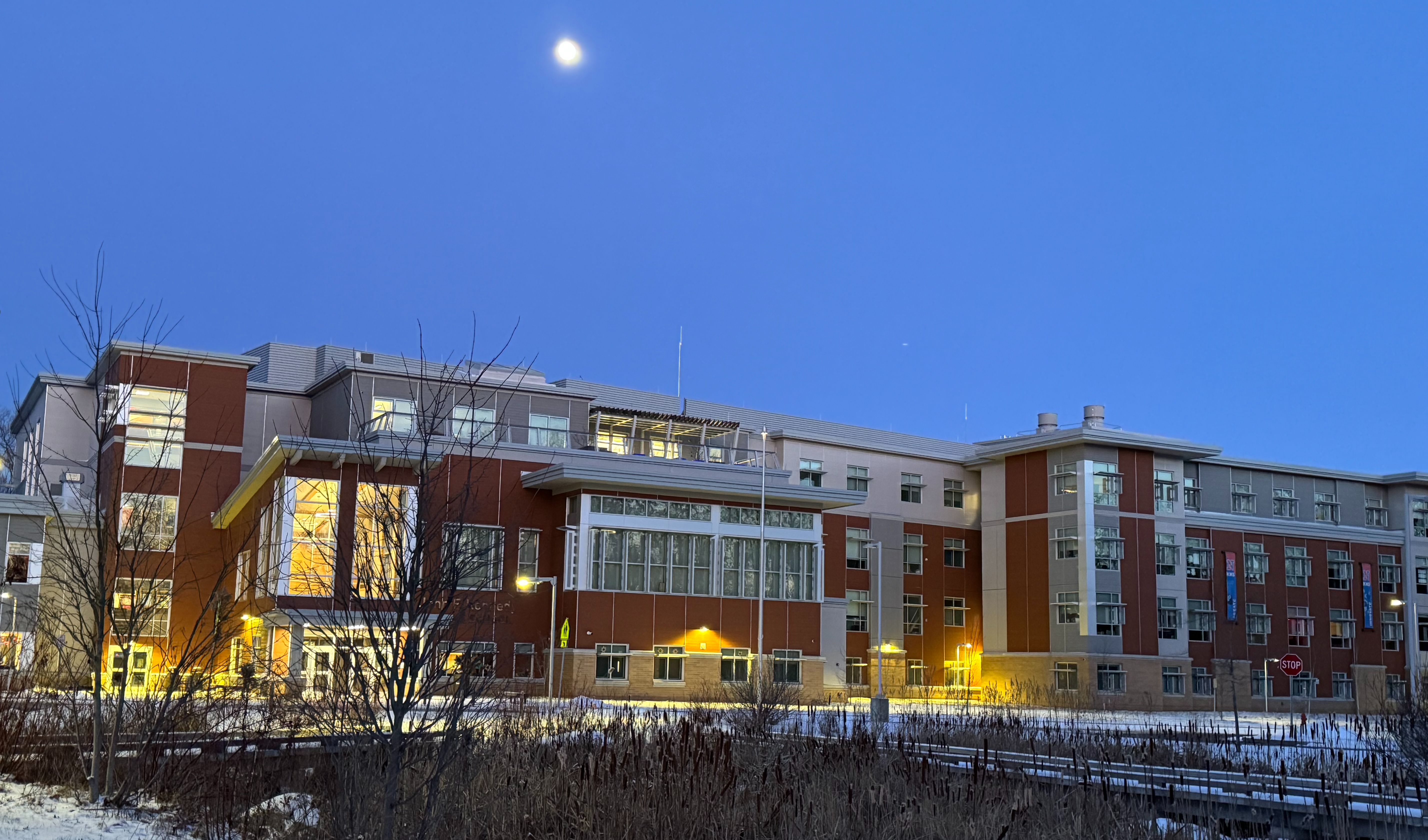 An image of the front of the building at dusk with a view of the moon and snow on the ground.