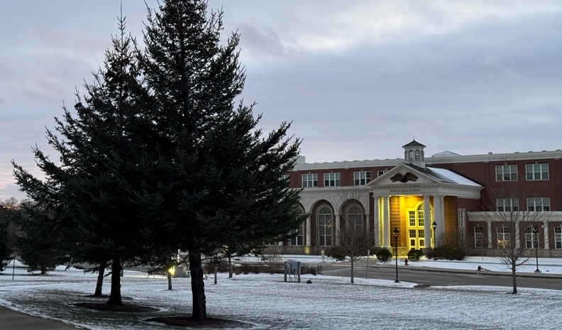 An image of the front of the building at dusk with a view of pine trees and snow.