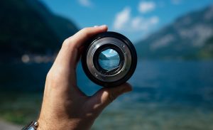 A hand holding a small, black camera lens, set against a serene outdoor backdrop with mountainous terrain and a blue sky.