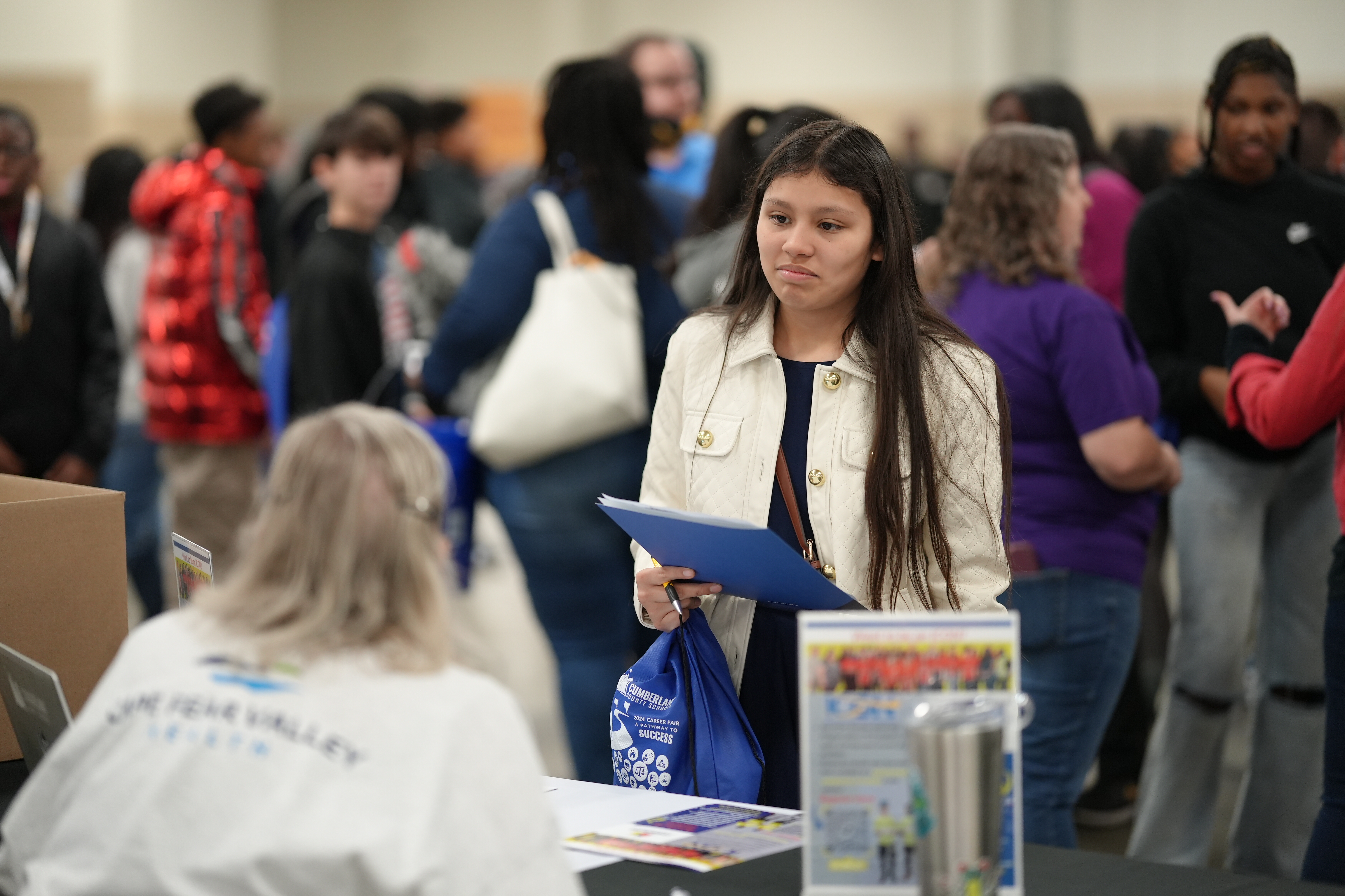 This image shows a young student standing at a booth during a career fair or educational event. She holds a blue folder and a branded drawstring bag, looking thoughtful as she interacts with someone seated at the booth, whose back is visible in the foreground. The booth has flyers and informational materials displayed, and several other students and adults are visible in the background, engaging in conversations and activities. The scene is busy, indicating a well-attended event.