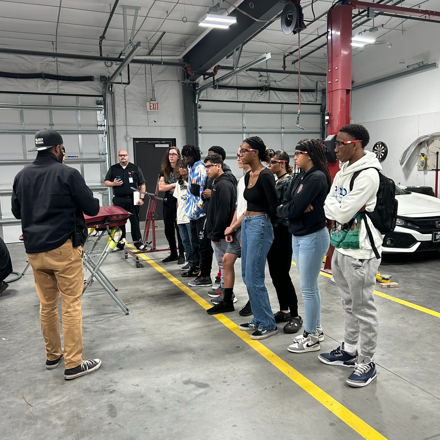 A group of students stands in a garage, intently listening to a man in a black cap and jacket who is leading a discussion on auto body repair. The students are lined up in front of a workbench, and in the background, another instructor watches the session. The setting is a spacious automotive workshop, with cars and repair equipment visible, providing a hands-on learning experience.