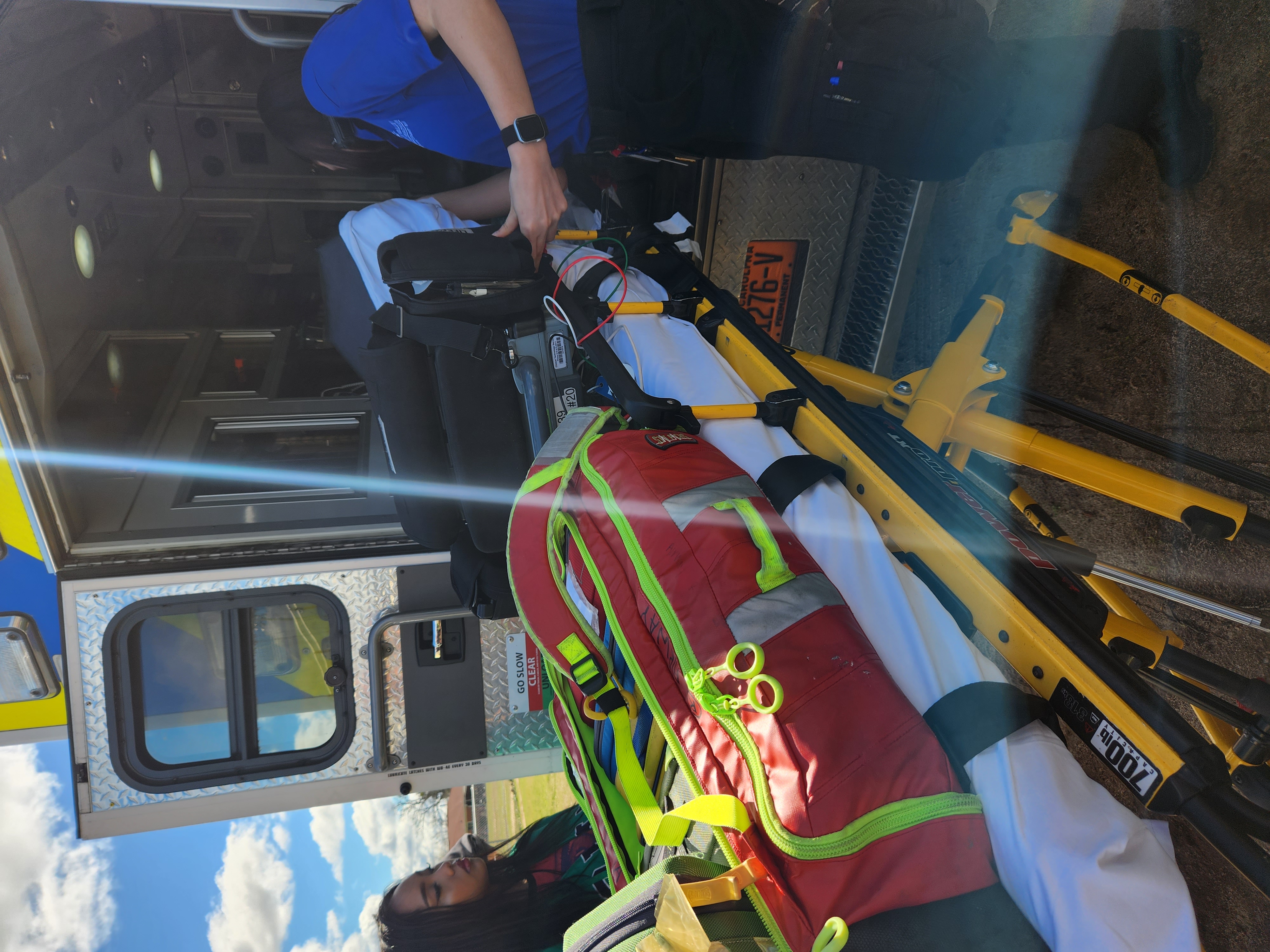 Am EMS professional is loading Medical equipment and a stretcher into  the back of an ambulance while a female student observes. 